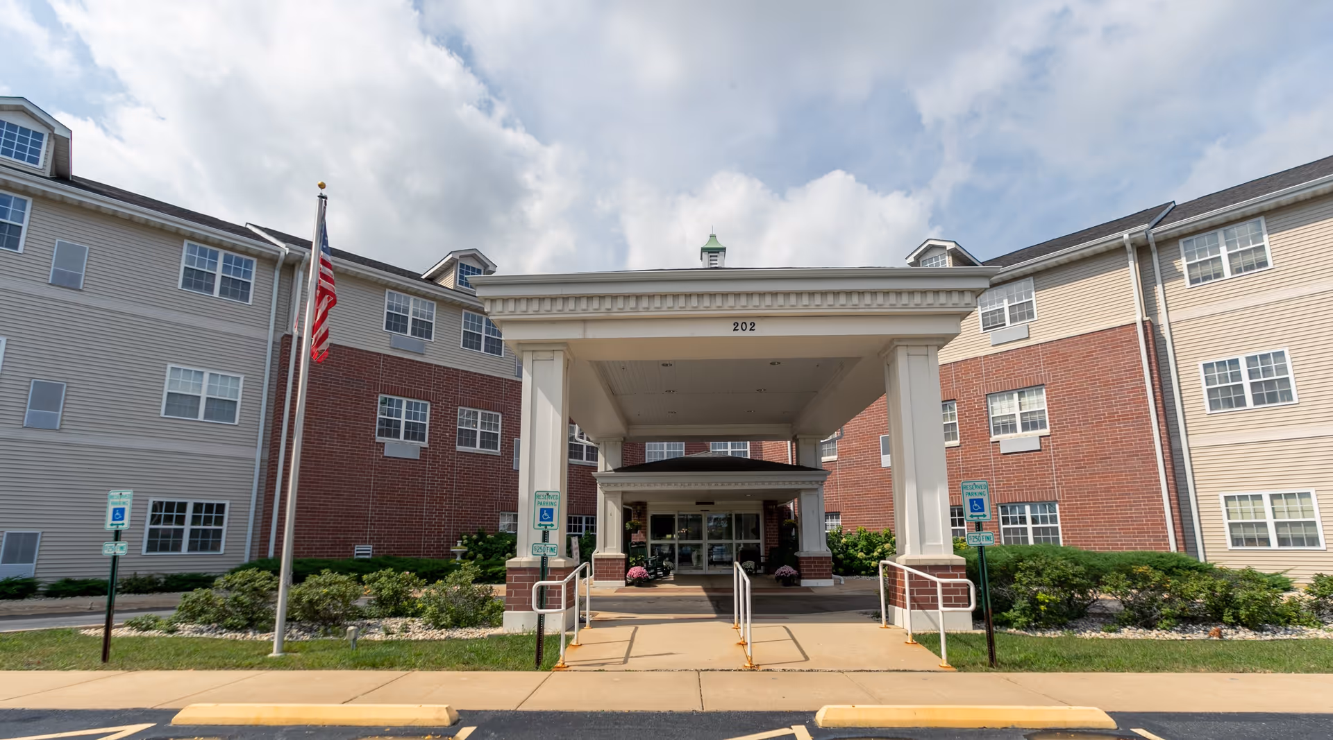 Front exterior view of Heritage Woods of Rockford building with a covered entrance, American flag, and reserved handicap parking signs on a partly cloudy day.