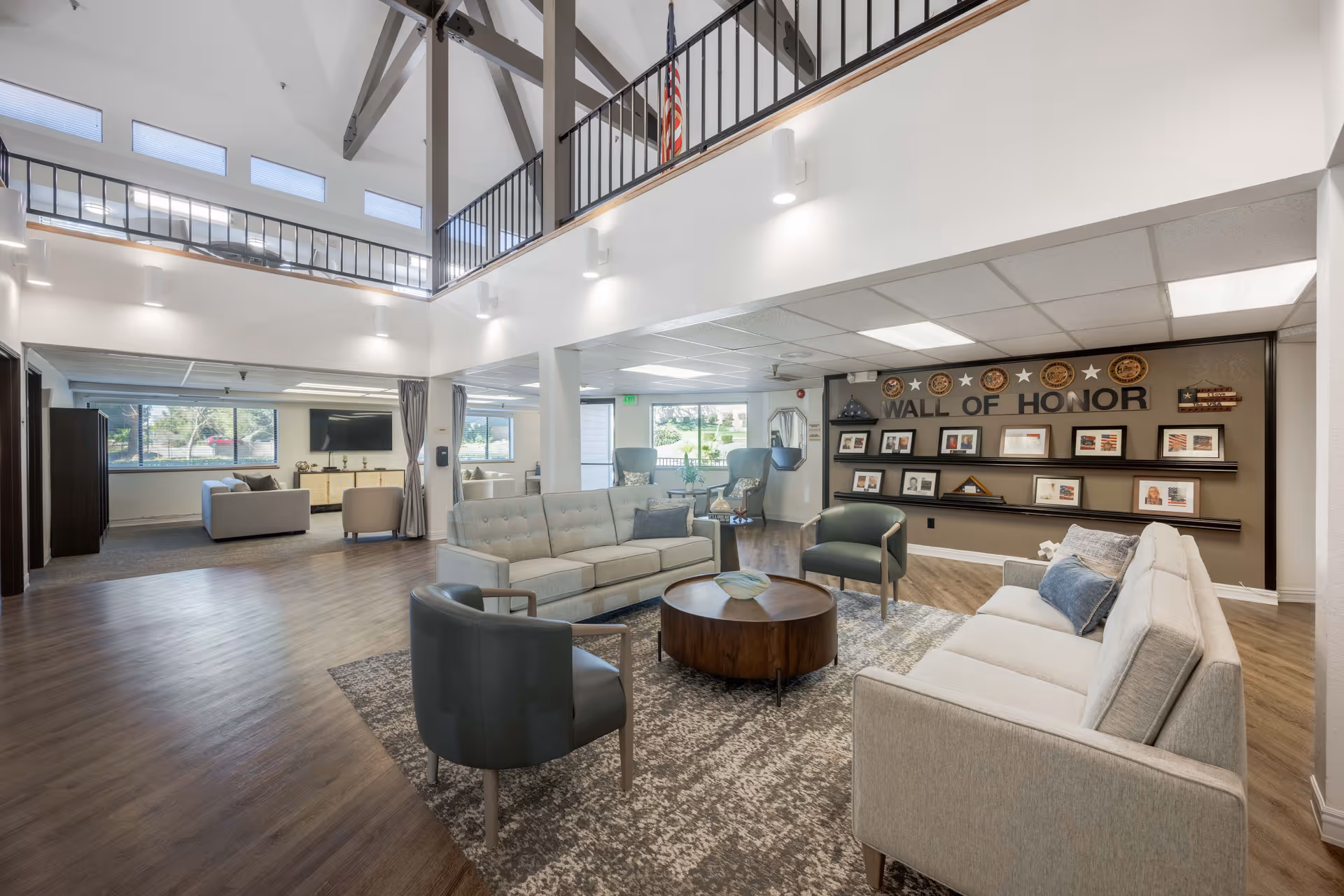 Spacious and well-lit senior living facility common area with modern furniture including sofas and chairs arranged around a circular wooden coffee table on a patterned rug. The room features high ceilings with exposed beams, large windows allowing natural light, and a 'Wall of Honor' displaying framed photos and plaques on the far wall.