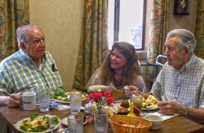 Two elderly men and a middle-aged woman sitting around a dining table with plates of food and drinks, engaged in conversation in a room with floral curtains and a window showing a parked car outside.
