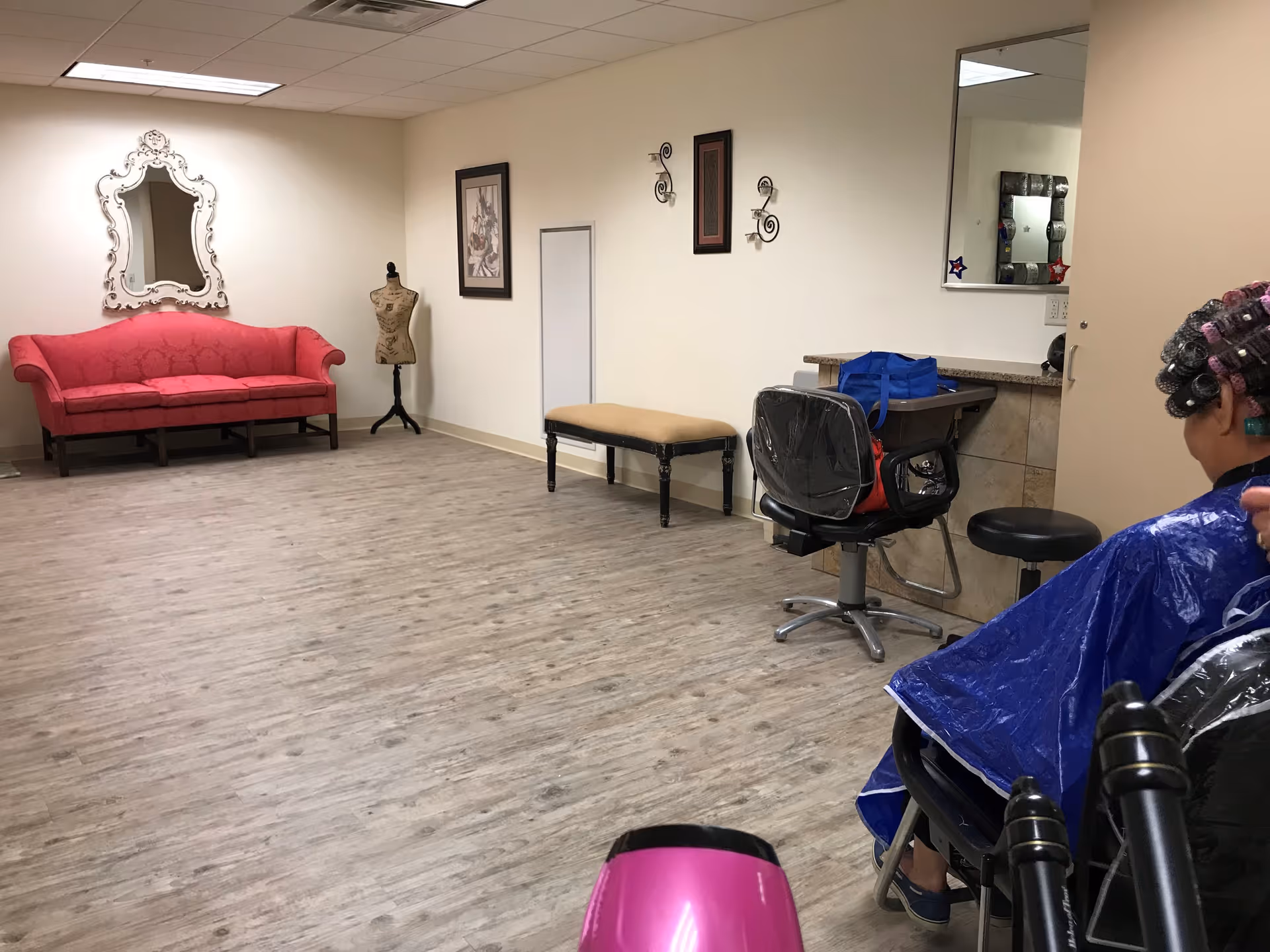 A spacious room with light wood flooring and cream-colored walls. On the left side, there is a bright pink upholstered sofa beneath a decorative white-framed mirror. Next to the sofa is a dress form mannequin. On the right side, there is a beige bench against the wall with two wall-mounted candle holders above it. A salon chair with a plastic cover is positioned in front of a counter with a mirror, where a person wearing a blue cape and hair rollers is seated. A pink hairdryer is visible in the foreground.