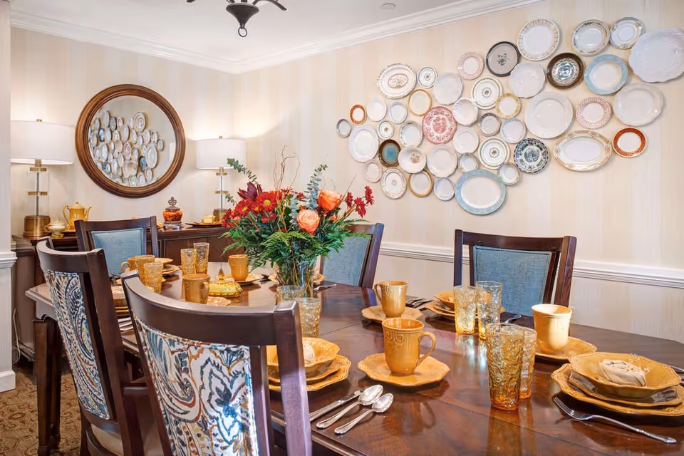 A dining room with a wooden table set for six people with yellow plates, mugs, and amber-colored glasses. There is a floral centerpiece with red and orange flowers in the middle of the table. The chairs have patterned upholstery on the backrests. On the wall, there is a decorative arrangement of various plates and a round mirror reflecting the plates. Two table lamps with white shades are on a sideboard against the wall.