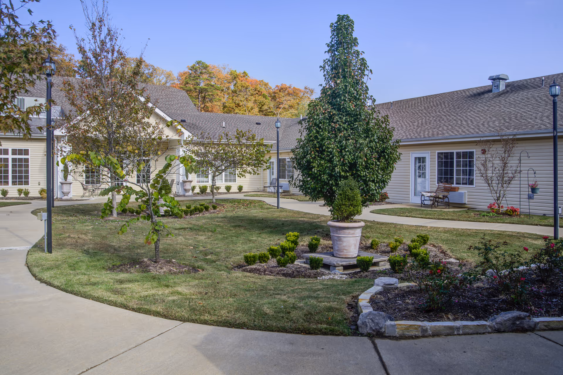 Outdoor courtyard area of Brookfield Assisted Living featuring a paved walkway surrounding a grassy area with small trees, shrubs, and a large potted plant in the center. The building with beige siding and multiple windows surrounds the courtyard under a clear blue sky.
