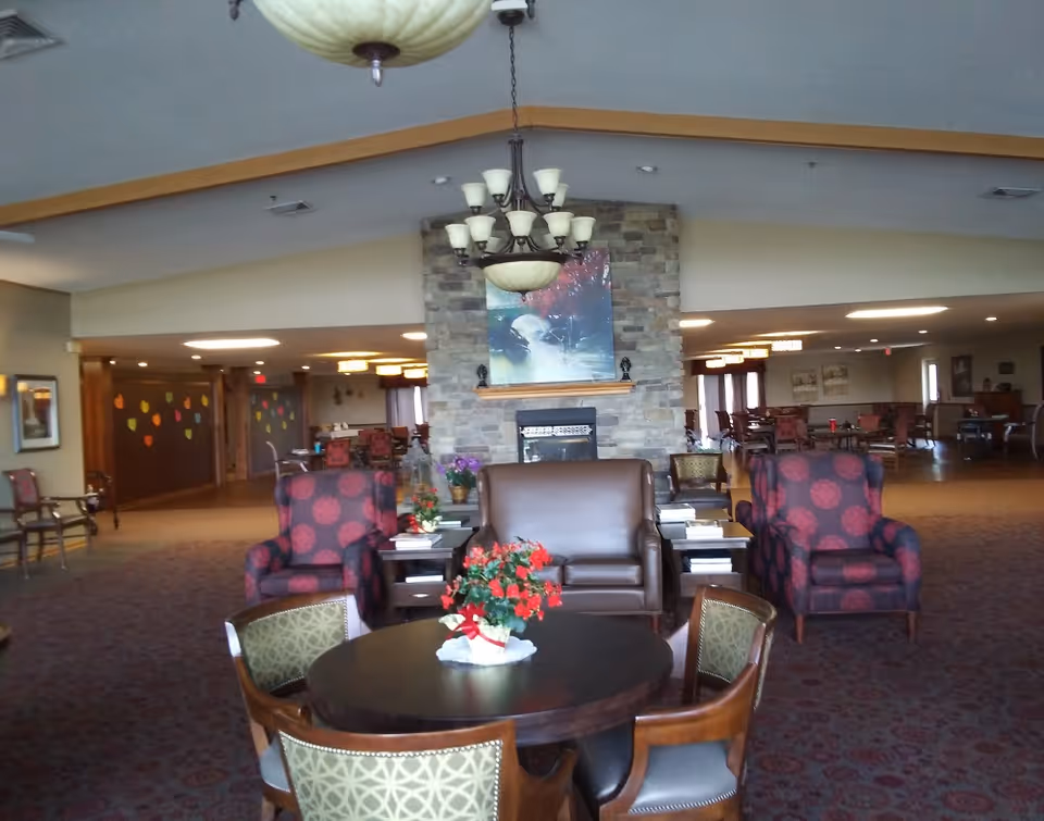 A spacious and well-lit common area in an assisted living facility featuring a stone fireplace with a painting above it, surrounded by comfortable seating including two patterned armchairs, a brown leather loveseat, and a round table with four chairs and a flower centerpiece. The room has carpeted floors, warm lighting, and additional seating and tables in the background.