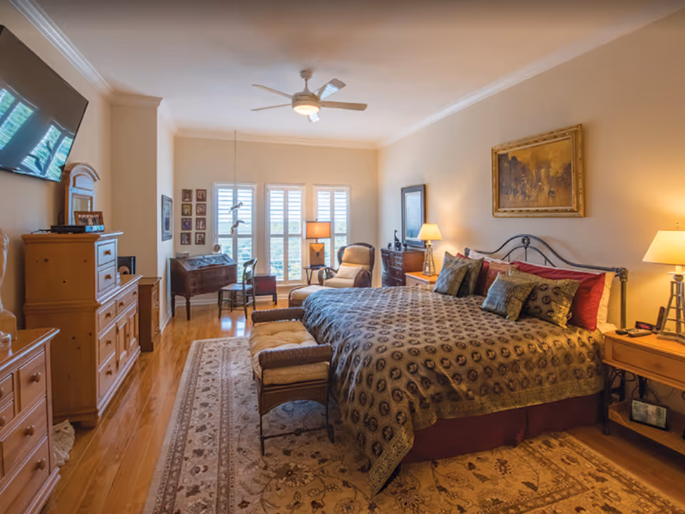 Spacious, well-lit bedroom with a large bed, patterned rug, dresser, seating area, and windows with shutters.