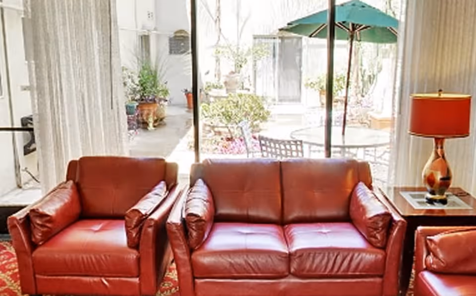 Red leather armchairs and a matching loveseat arranged before a sliding glass door overlooking a patio with a table and green umbrella.