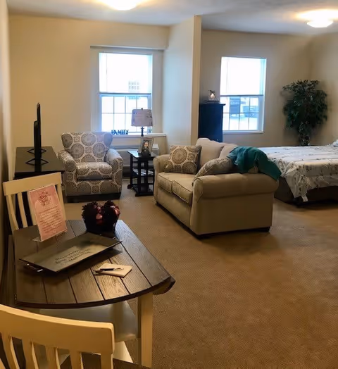 A cozy studio apartment with a beige sofa, patterned armchair, wooden coffee table, and a bed in the background. The room has two windows letting in natural light, a small side table with a lamp, and a potted plant in the corner.