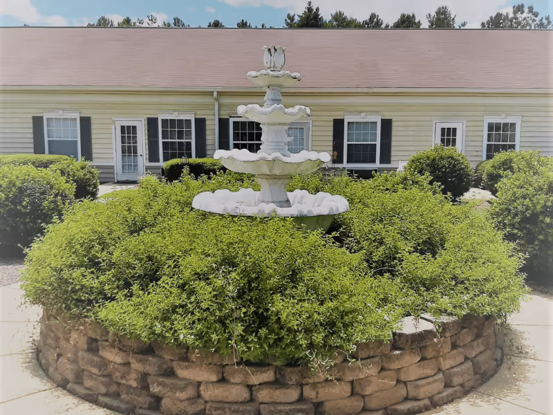 A white multi-tiered fountain surrounded by green bushes and a circular stone wall in front of a single-story building with beige siding, black shutters, and several windows and doors under a clear blue sky.