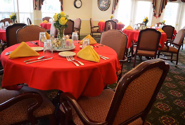 A dining room with round tables covered in red tablecloths, each set with yellow folded napkins, plates, silverware, and glasses. The chairs around the tables have patterned upholstery. The room is well-lit with natural light coming through the windows, and there are floral centerpieces on each table.