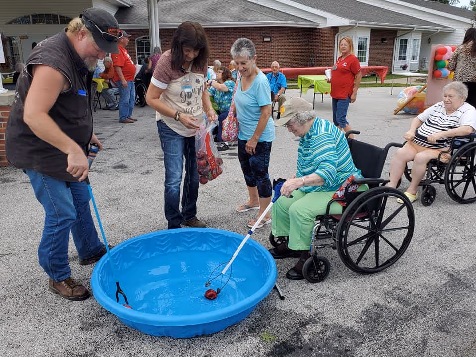 A group of elderly people and caregivers are gathered outside on a paved area near a building. Two elderly women in wheelchairs are playing a game involving fishing for floating objects in a small blue plastic pool filled with water, using long-handled nets. Other people stand nearby watching and assisting. The setting appears to be a community event at a senior living facility.