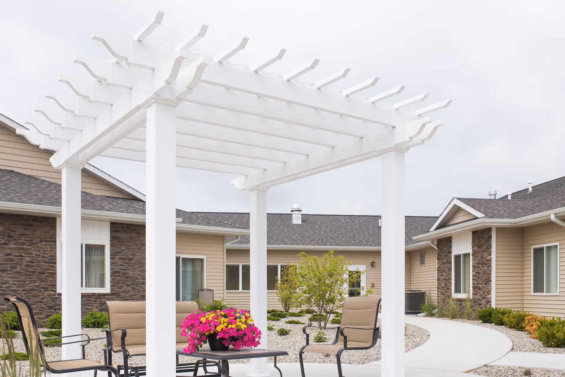 White pergola over patio chairs and a table with a bright flower pot in a landscaped courtyard outside single-story senior living buildings.
