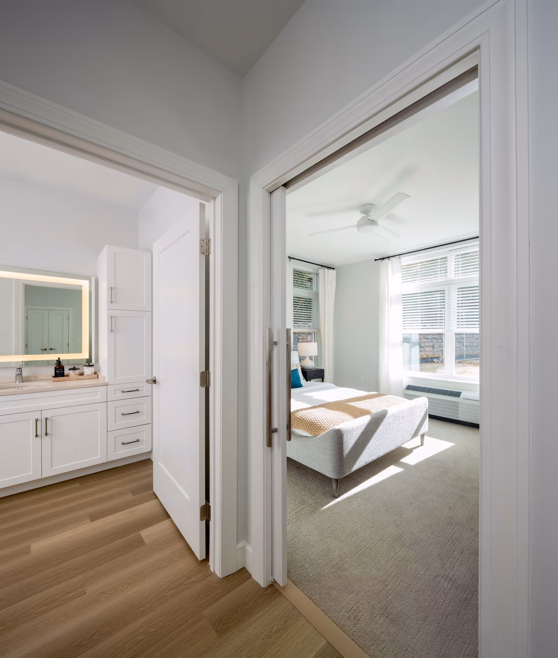 View of a bright bedroom with a bed, bedside table with lamp, and large windows with white curtains, adjacent to a bathroom with white cabinetry, a sink, and a lighted mirror. Both rooms have light-colored walls and flooring.