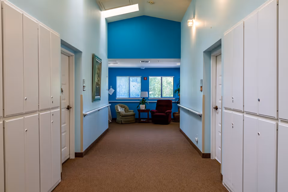 A hallway in Oakview Village Independent Senior Living with light blue walls, white doors, and white storage cabinets on both sides. At the end of the hallway, there are two armchairs, one green and one red, with a small table and a plant between them, positioned in front of two windows. The ceiling is vaulted with a skylight, and there is a framed painting on the left wall.