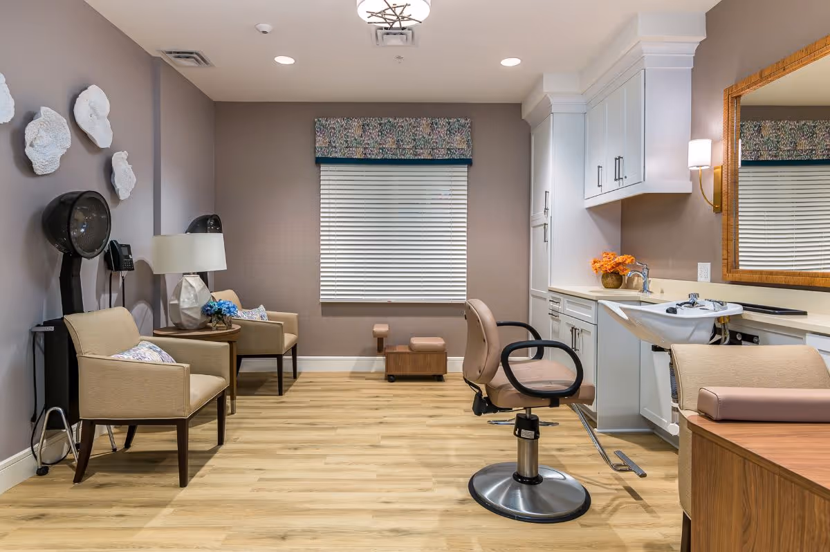 A well-lit salon area in a senior living facility featuring a styling chair in front of a wash basin, two beige armchairs with decorative pillows, a side table with a lamp and flowers, wall-mounted hair dryers, and a large mirror above a countertop with cabinets. The room has light wood flooring, neutral-colored walls, and a window with blinds and a patterned valance.