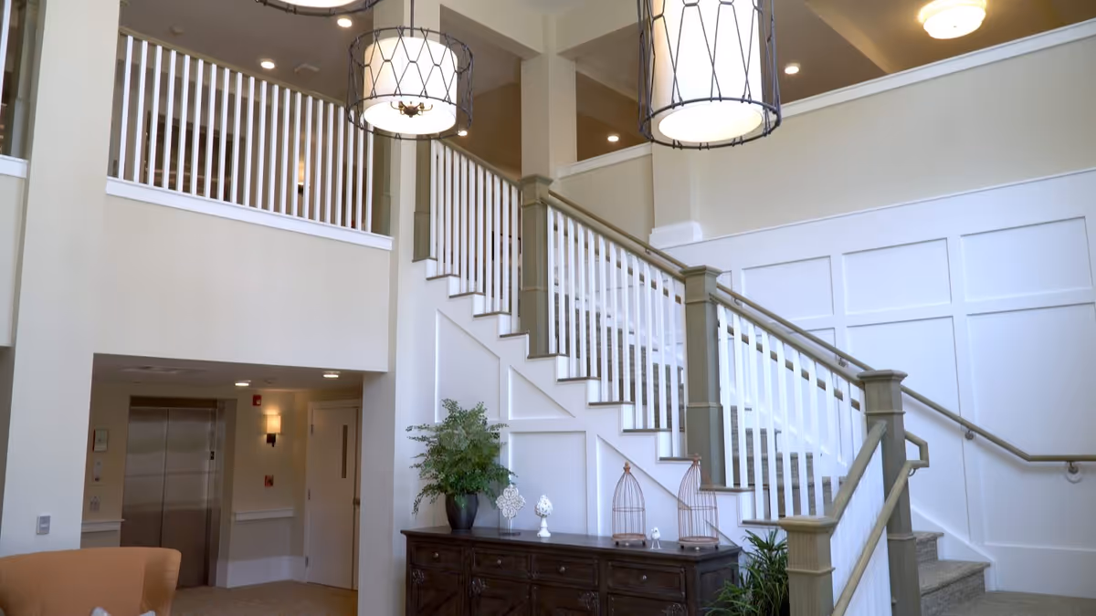 Interior view of a senior living facility showing a staircase with white railings and beige carpeting. Below the staircase is a dark wooden cabinet decorated with plants and ornamental items. Two large hanging light fixtures are visible above, and an elevator and door are seen in the background.