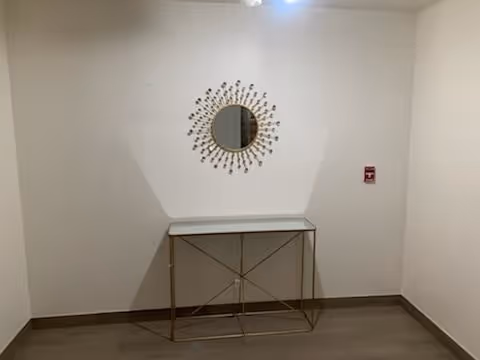 A small corner of a room with a decorative round mirror on the wall and a narrow console table with a glass top and gold-colored metal frame below it. The walls are white, and there is a fire alarm pull station mounted on the right wall.
