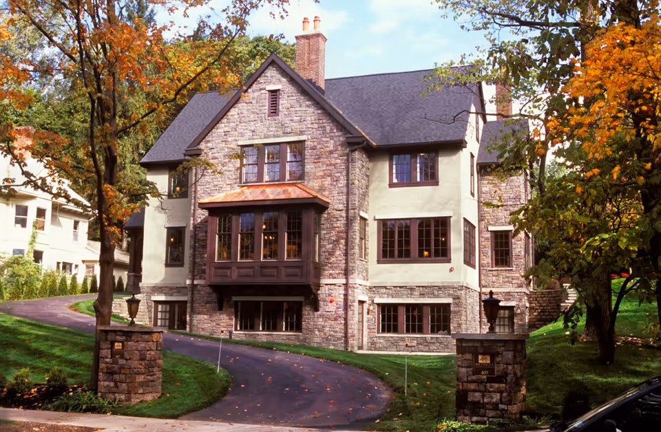 A large, elegant stone and stucco building with multiple windows and a steep roof, surrounded by green lawns and trees with autumn foliage. A curved driveway leads up to the building, flanked by stone pillars.