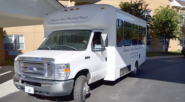 A white shuttle bus parked outside a building under a covered driveway. The bus has the text 'Schroder Lane Church of Christ' written on the front and side. The building in the background is beige with windows and some trees nearby.