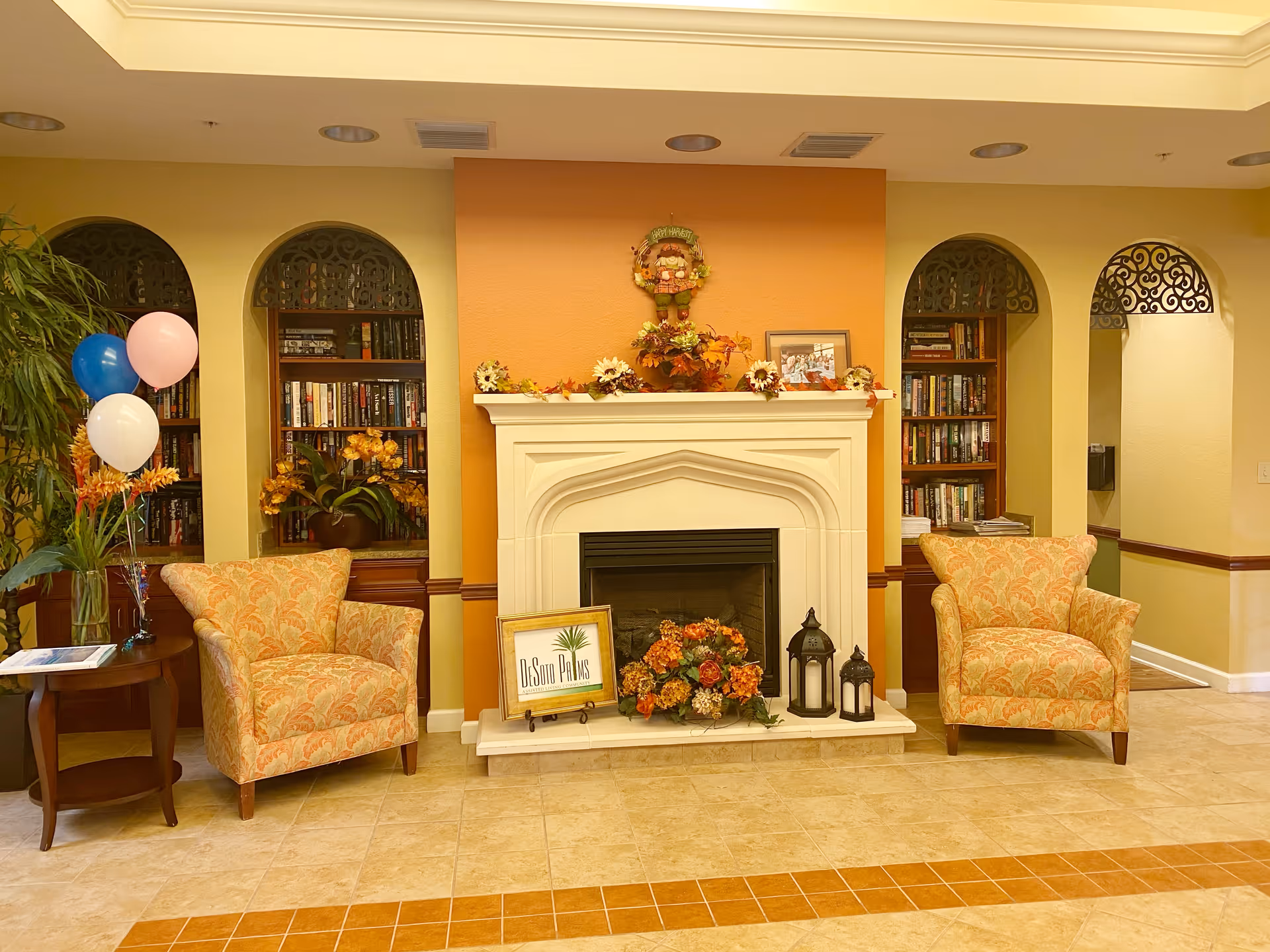 A cozy sitting area in DeSoto Palms Assisted Living Community featuring two patterned armchairs on either side of a white fireplace decorated with autumn-themed flowers and lanterns. Behind the chairs are built-in bookshelves filled with books, and a small table with balloons is placed next to the left chair. The walls are painted in warm tones of yellow and orange.