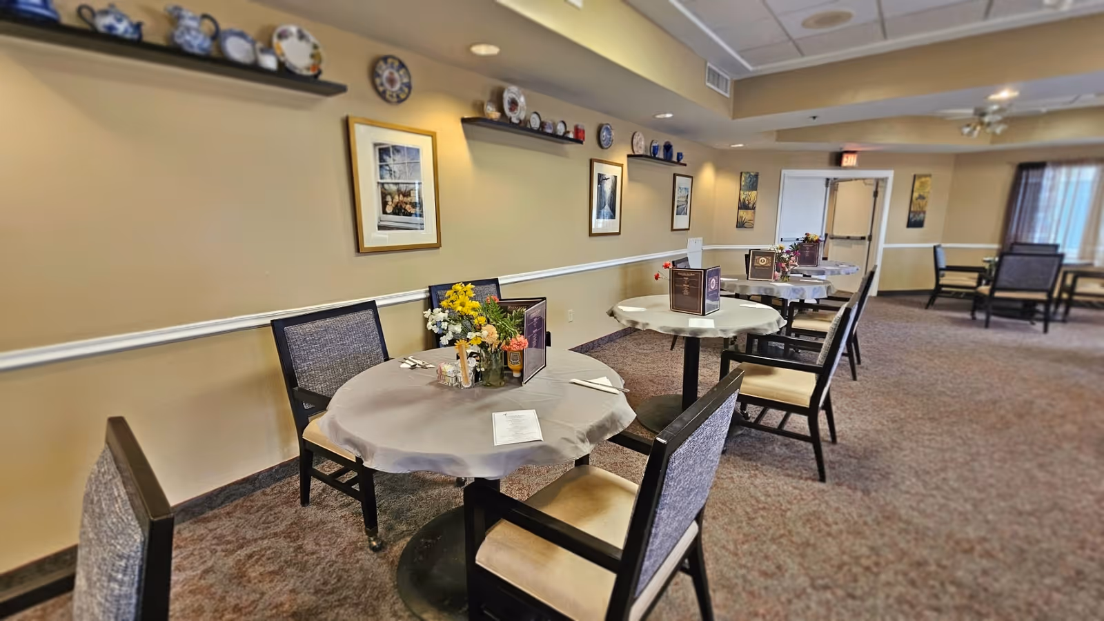 Dining room with round tables, chairs, floral centerpieces and decorative plates on wall shelves.
