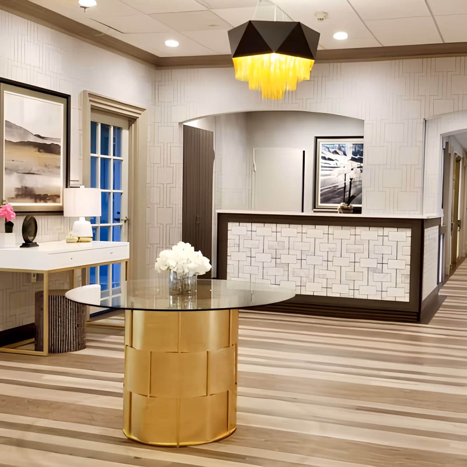 A modern reception area with a gold cylindrical table topped with a glass surface and a vase of white flowers. Behind the table is a reception desk with a patterned front and a framed black and white photograph on the wall. To the left, there is a white console table with a lamp and decorative items, and a framed abstract painting above it. The floor has a striped wood pattern, and a unique black and yellow ceiling light fixture hangs above.