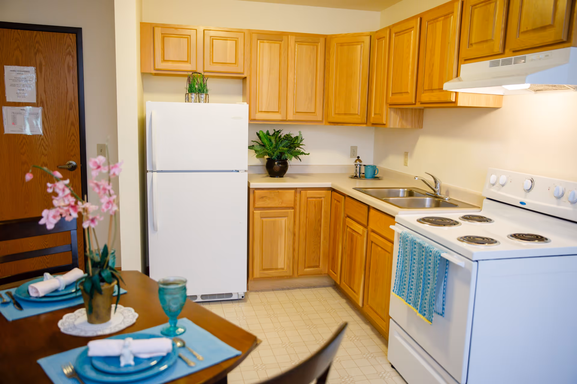 A clean and well-lit kitchen with wooden cabinets, a white refrigerator, a white electric stove with a blue towel hanging on the handle, and a double sink. In the foreground, there is a dining table set with blue placemats, plates, rolled napkins, silverware, a green glass, and a potted pink orchid. A wooden door with papers taped to it is visible in the background.
