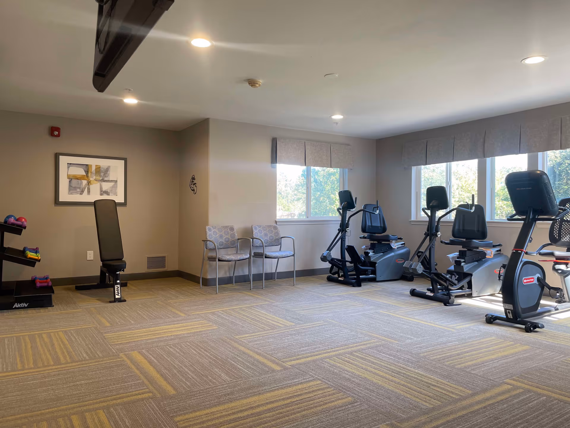 A fitness room with exercise equipment including stationary bikes, a weight bench, and a rack of dumbbells. Two chairs are placed against the wall under a window with natural light coming in. The room has patterned carpet flooring and neutral-colored walls with a framed abstract artwork.
