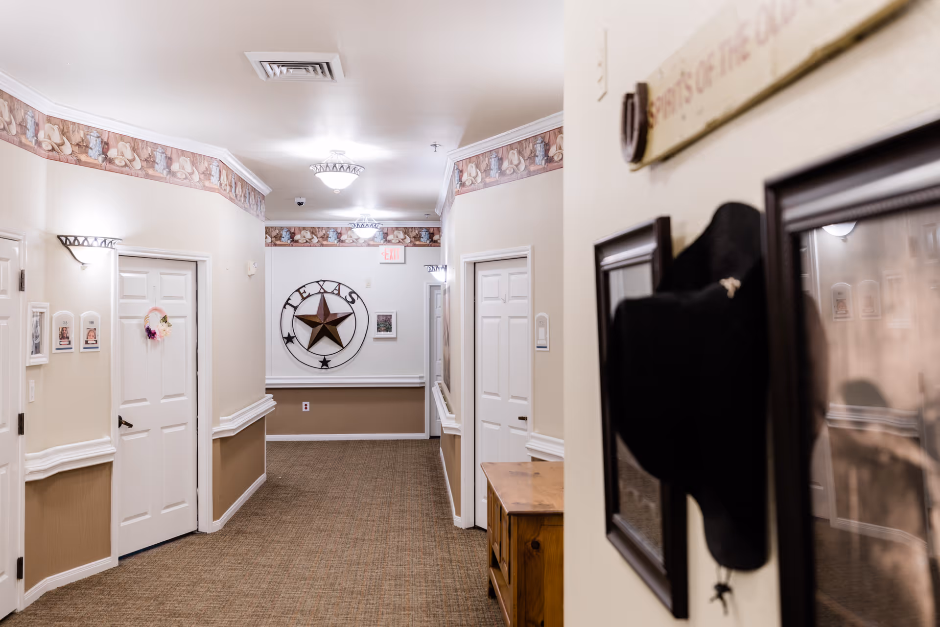A well-lit hallway in a senior living facility with beige walls and carpeted floor. The hallway features white doors on both sides, decorative wall trim, and a large Texas star emblem on the far wall. There are framed pictures and a black cowboy hat hanging on the right wall. The ceiling has recessed lighting and a decorative border near the ceiling.