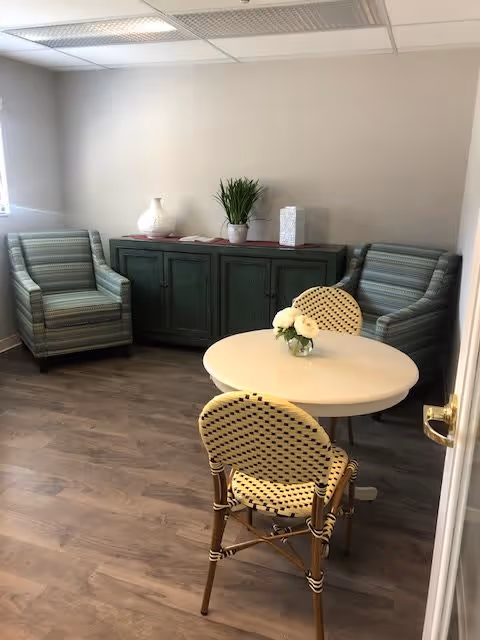 A small sitting area with two striped armchairs positioned against a wall with a green cabinet between them. In front of the chairs is a round white table with two woven chairs and a small vase with white flowers on top. The room has light-colored walls and wood flooring.