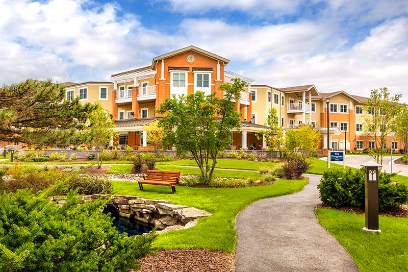 A large, multi-story assisted living facility building with a well-maintained garden in the foreground. The garden features green grass, trees, shrubs, a small stone bridge over a water feature, a wooden bench, and a paved walking path leading to the building entrance. The sky is partly cloudy with blue patches.