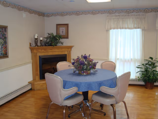 A small dining area with a round table covered with a blue tablecloth and four beige chairs around it. A floral centerpiece is on the table. In the background, there is a wooden fireplace with decorative candles and a plant on the mantel. A window with sheer white curtains lets in natural light, and a potted plant is placed near the window. The room has light-colored walls and wooden flooring.