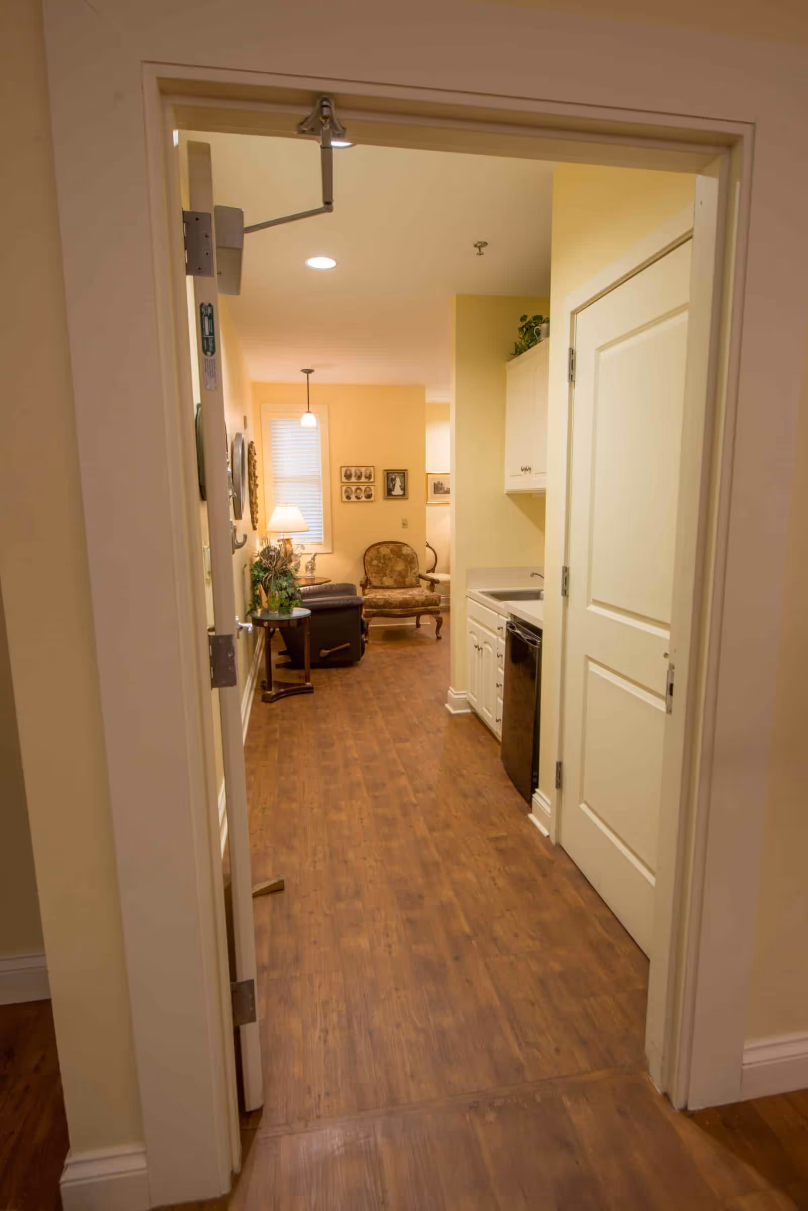 View through an open doorway into a hallway with wooden flooring leading to a small living area. The living area has a patterned armchair, a side table with a lamp, and framed pictures on the wall. To the right of the hallway is a small kitchenette with white cabinets, a sink, and a dishwasher.