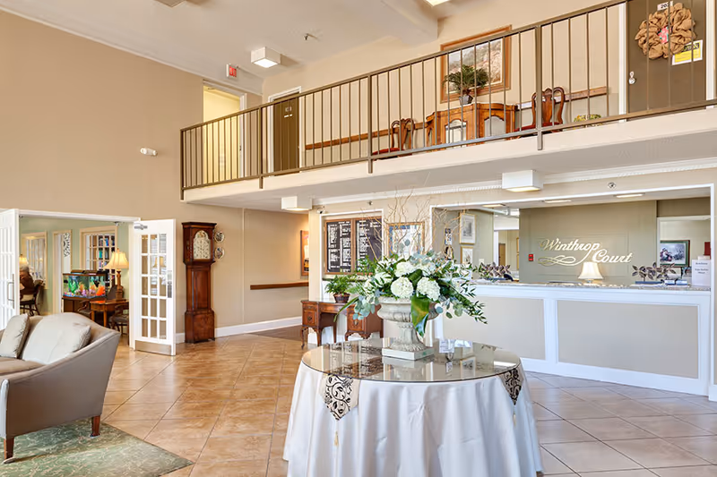 Bright two-story reception lobby of Winthrop Court with a round table floral centerpiece, seating area, and front desk.