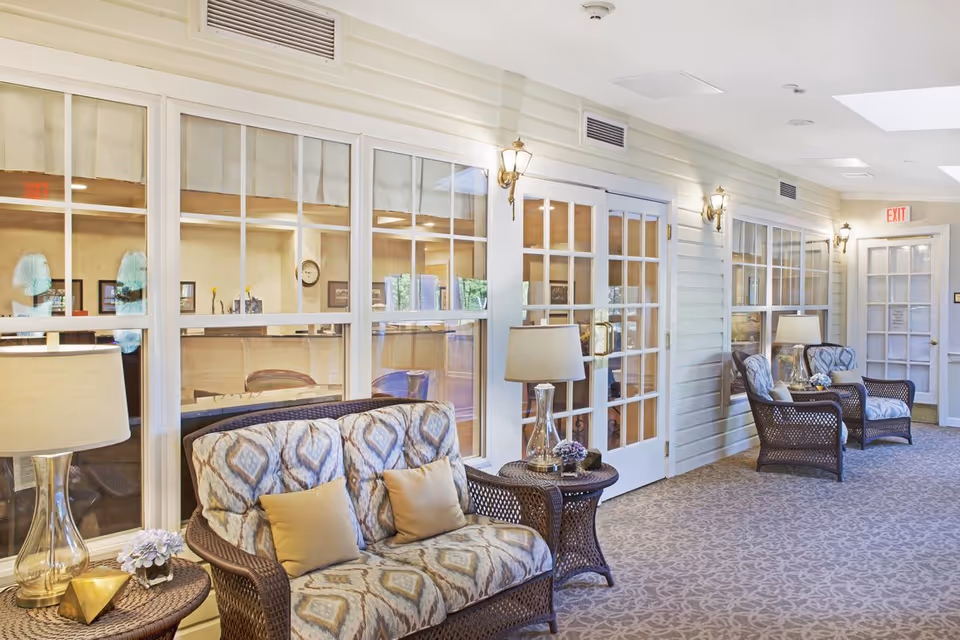 Bright corridor lounge with patterned wicker chairs, side tables and lamps along glass-paneled doors.