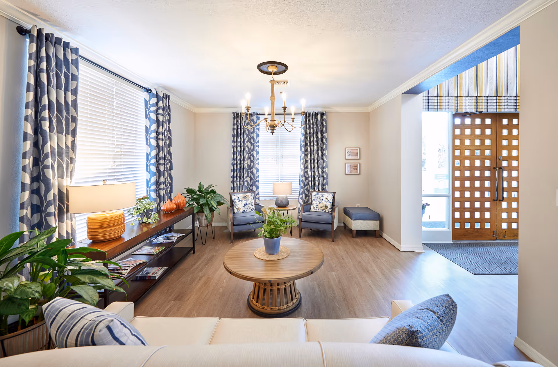 A bright and cozy living room area with a white couch adorned with blue and white pillows in the foreground. A round wooden coffee table with a potted plant sits in the center of the room. Two blue armchairs with floral cushions are placed against the back wall, flanked by windows with blue and white patterned curtains. A wooden console table with a lamp, plants, and magazines is along the left wall. The room has light-colored walls, wood flooring, and a chandelier hanging from the ceiling. To the right, there is a wooden double door entrance with a striped valance above it.