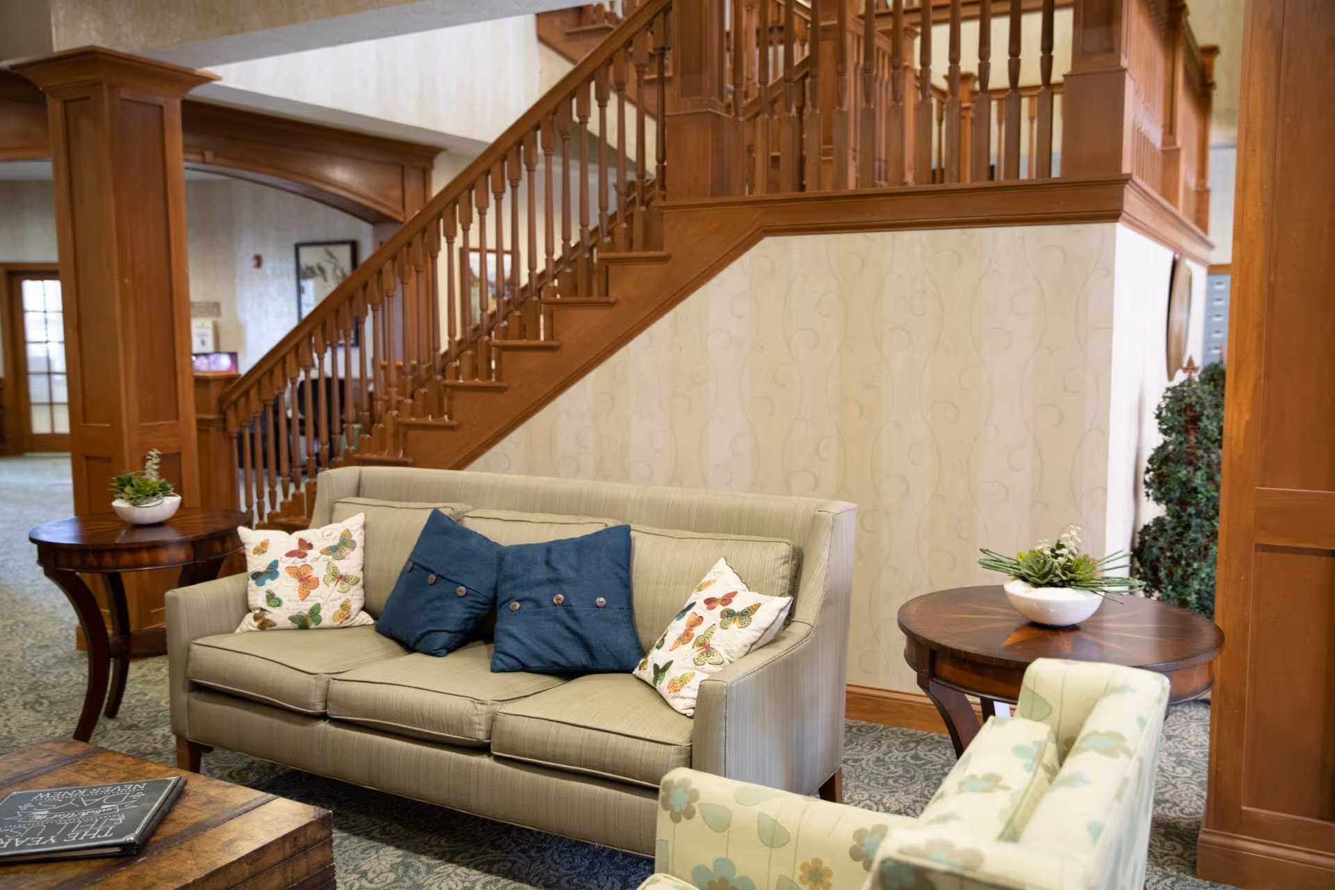 A cozy seating area in a senior living facility with a beige sofa adorned with butterfly and blue buttoned pillows, a floral patterned armchair, two wooden side tables with small potted plants, and a wooden staircase with a railing in the background.