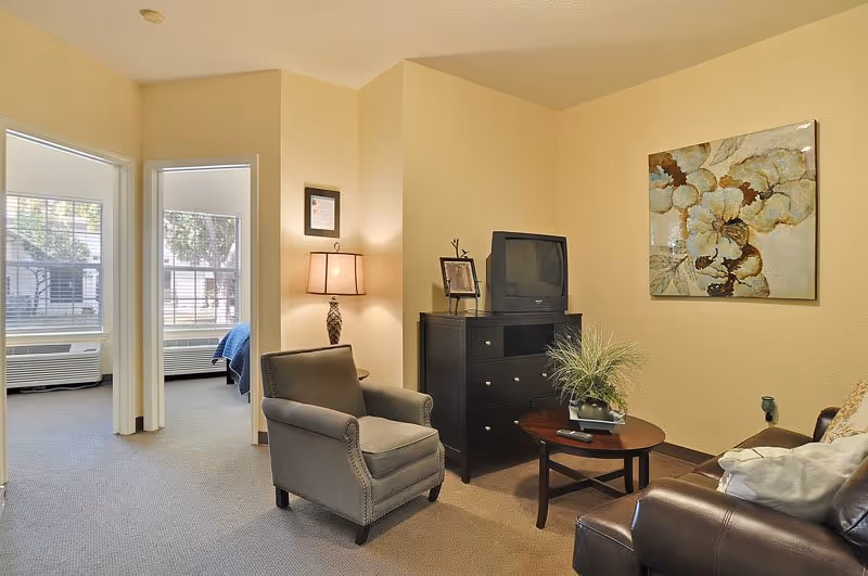 A cozy living room area in a senior living facility with a gray armchair, a dark wooden TV stand holding an older model television, a small round coffee table with a plant, and a brown leather sofa. The walls are painted light beige and decorated with a large floral painting. Two doorways lead to bedrooms with large windows and blue bedding visible.