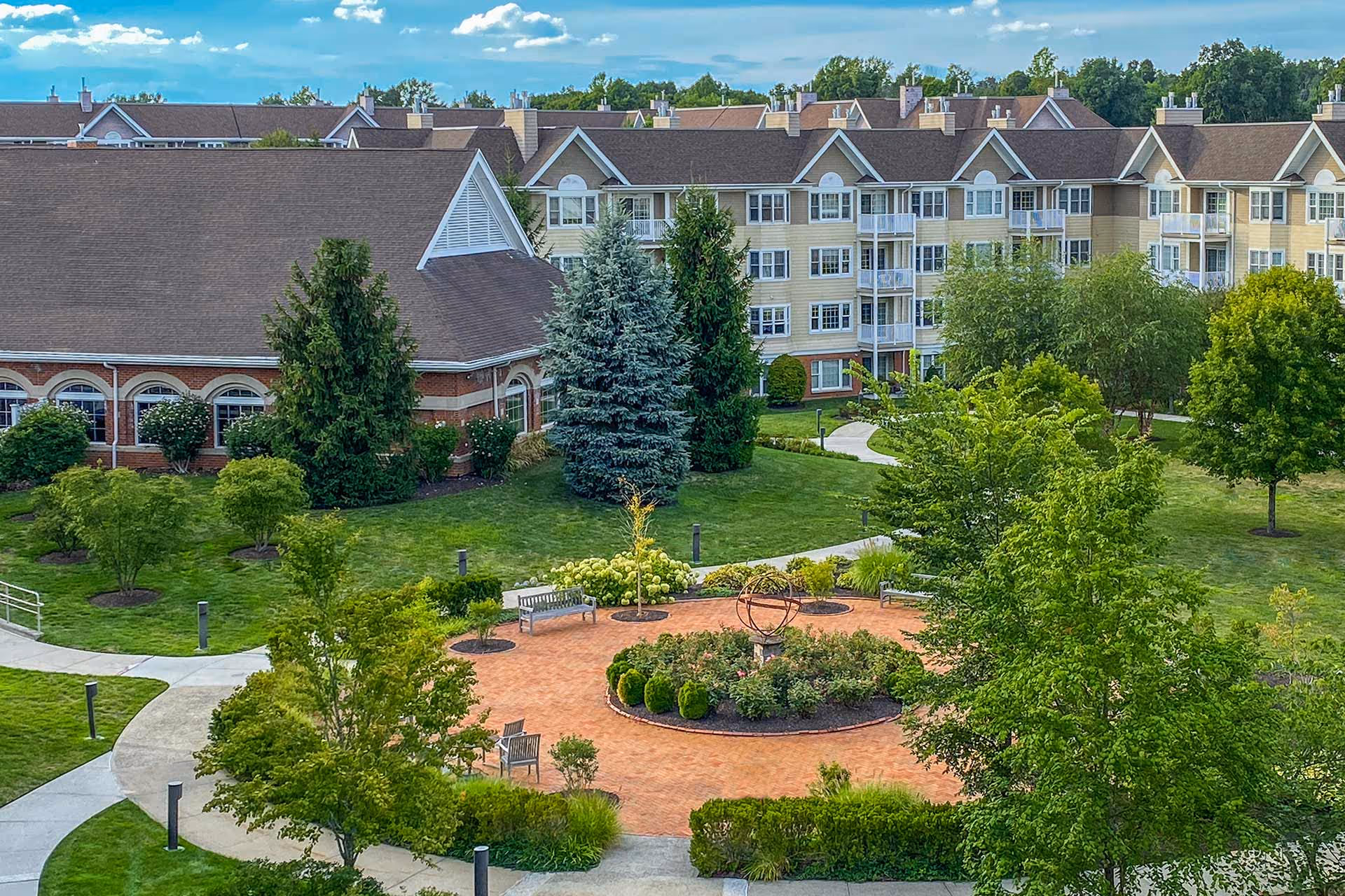 A landscaped outdoor garden area with a circular brick patio featuring a decorative metal sculpture in the center, surrounded by benches and greenery. In the background, there is a multi-story residential building and a large building with a steep roof and arched windows, under a partly cloudy blue sky.