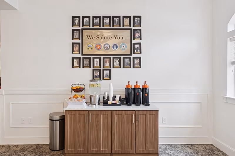 A refreshment station with a wooden cabinet holding a fruit basket, a beverage dispenser with lemon water, disposable cups, and three black coffee dispensers. Above the station is a wall display with framed photographs surrounding a sign that reads 'We Salute You...' along with military service emblems.