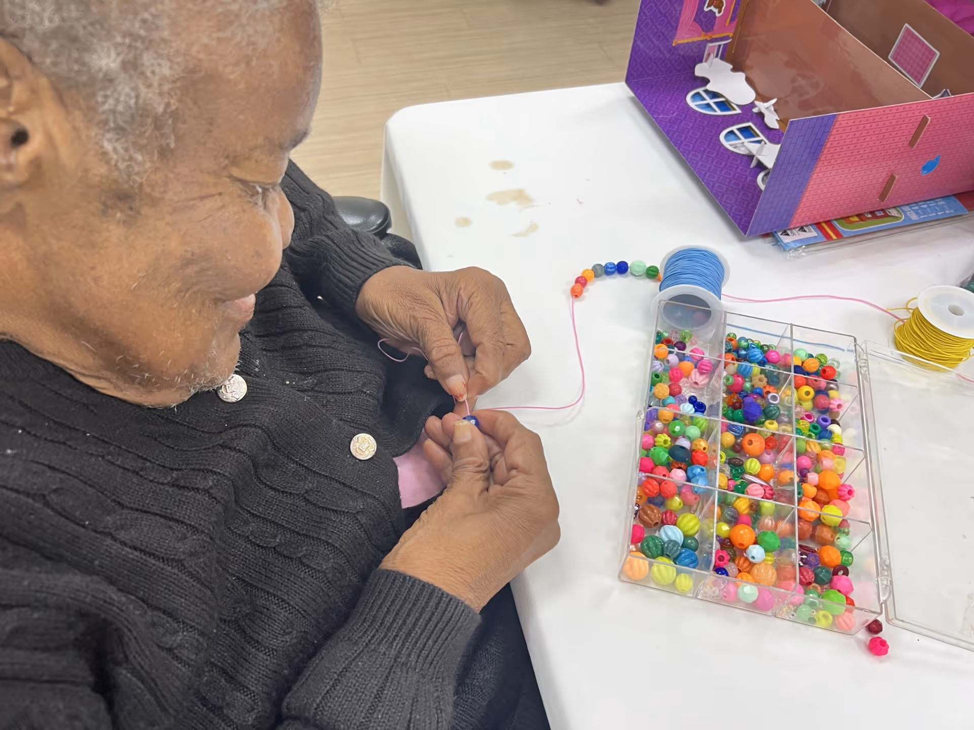 An elderly person wearing a black sweater is threading colorful beads onto a pink string at a white table. A clear plastic organizer filled with various colorful beads and spools of string are also on the table.
