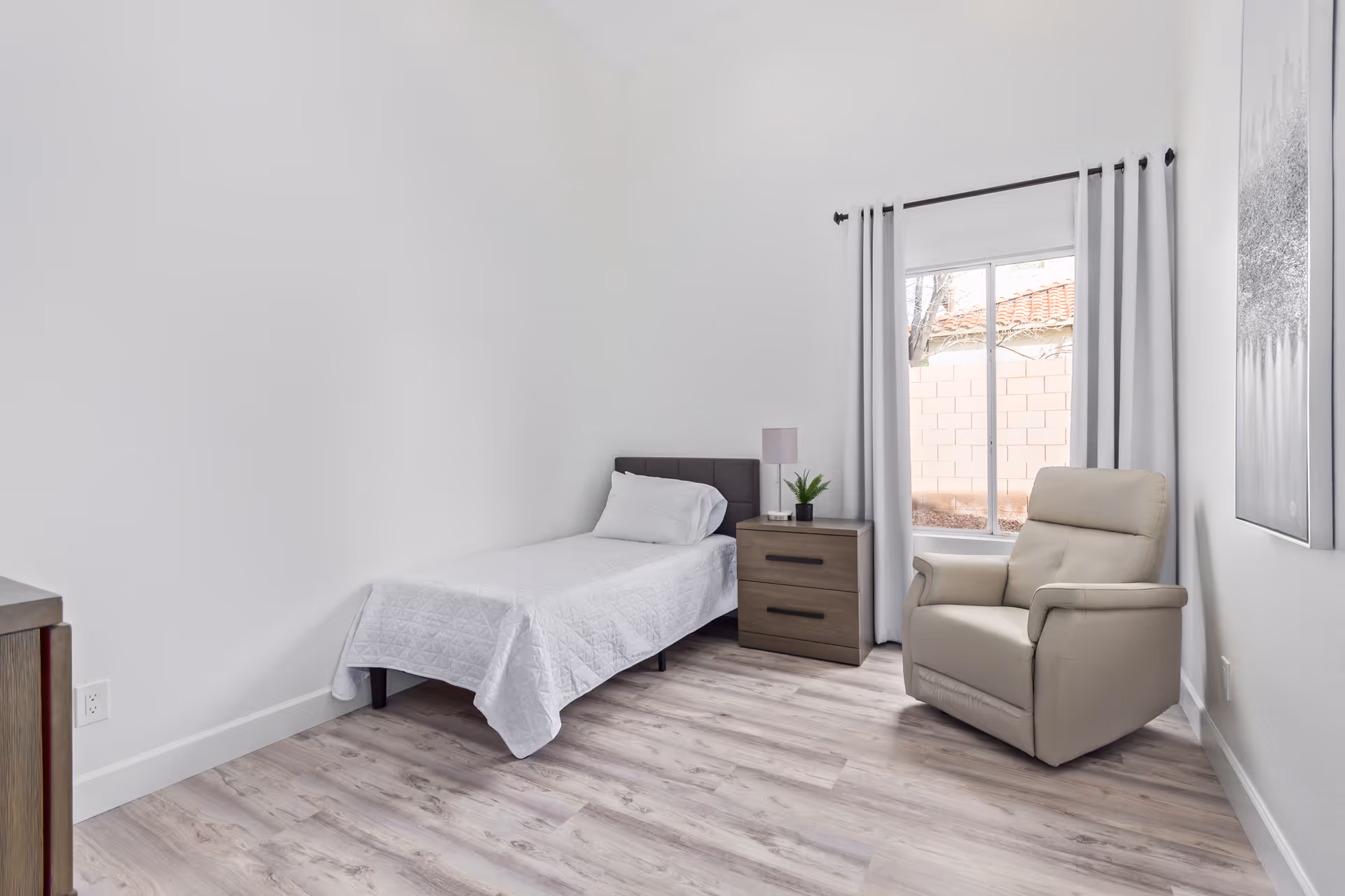 A simple, clean bedroom with a single bed covered in a white quilt, a wooden nightstand with a lamp and small plant, a beige armchair, a large window with white curtains, and light wood flooring.