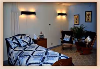 A senior care facility bedroom with a blue-and-white quilted bed, nightstand, chairs, and plants.