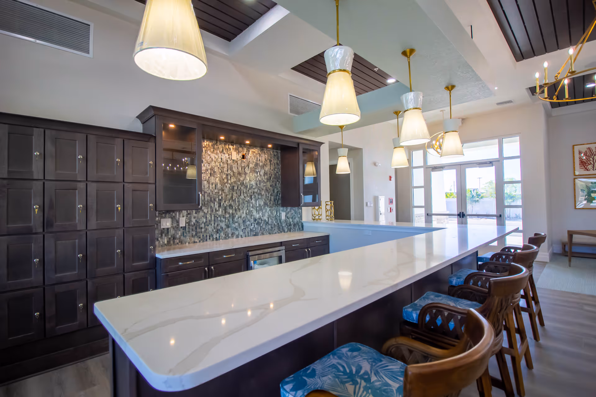A modern kitchen area with a long white marble countertop island surrounded by wooden bar stools with blue patterned cushions. The back wall features dark wood cabinets and a mosaic tile backsplash. Several pendant lights hang from the ceiling, illuminating the space. Large windows and glass doors allow natural light to enter the room.