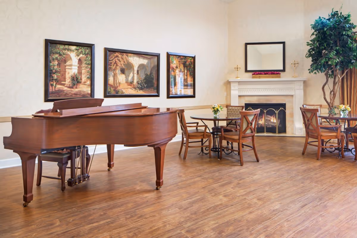 A spacious room with a wooden grand piano on the left, three framed paintings on the wall behind it, and a cozy fireplace with a mirror above it on the right. There are round tables with wooden chairs around them, small flower arrangements on the tables, and a large potted plant near the fireplace. The floor is wooden and the walls are light-colored with subtle patterns.