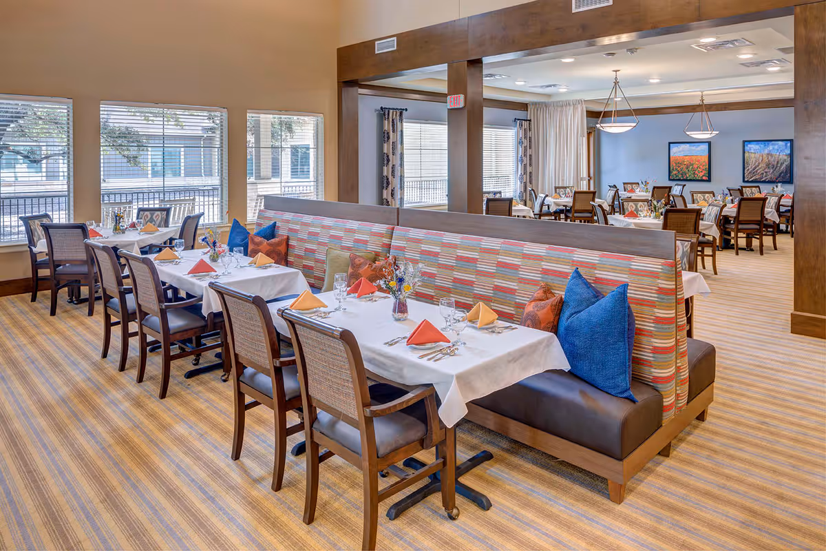 A bright and spacious dining room with multiple tables set for a meal. The tables have white tablecloths, colorful folded napkins, glassware, and small floral centerpieces. There is a long booth with a striped backrest and blue and orange pillows. Large windows allow natural light to fill the room, and framed artwork hangs on the walls.