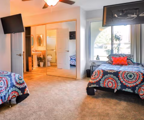 Spacious bedroom with two twin beds in colorful patterned bedding, mirrored closet doors, a window, wall-mounted TV, and an en-suite bathroom visible.