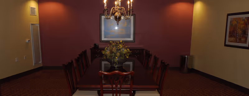 A formal dining room with a long wooden table surrounded by chairs. A chandelier hangs above the table, and a floral centerpiece is placed in the middle. The walls are painted in warm tones with framed artwork hanging on them.