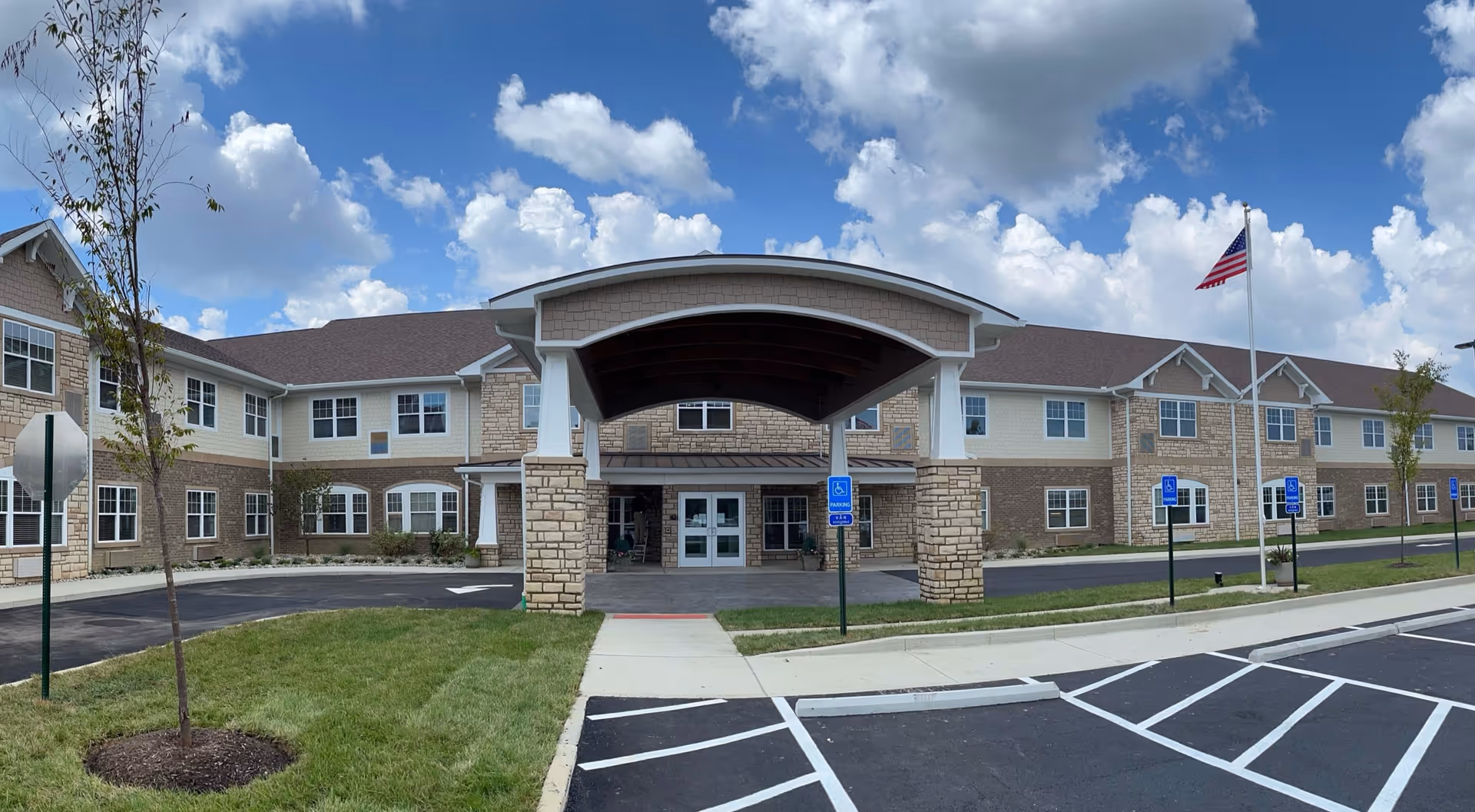 Front entrance of a two-story senior living facility with a covered porte-cochère, flagpole, and marked parking spaces under a partly cloudy sky.