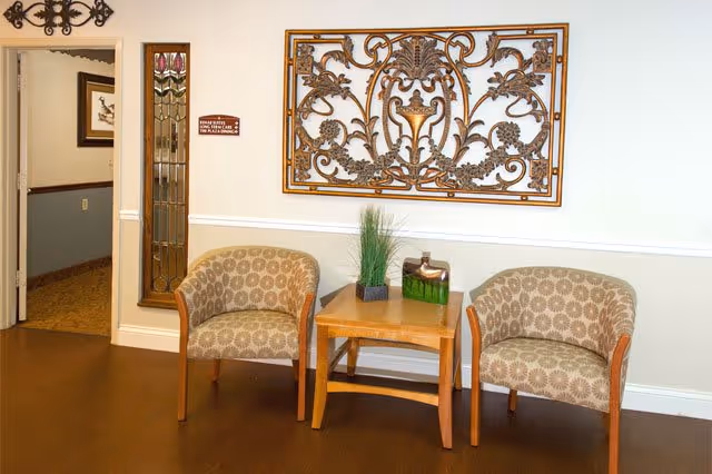 A small seating area with two patterned armchairs and a wooden side table between them. On the table are decorative items including a small plant and a green glass container. Above the chairs is an ornate metal wall decoration. To the left, there is a doorway with a stained glass panel and a sign indicating Broadway Rehabilitation and Care Center.