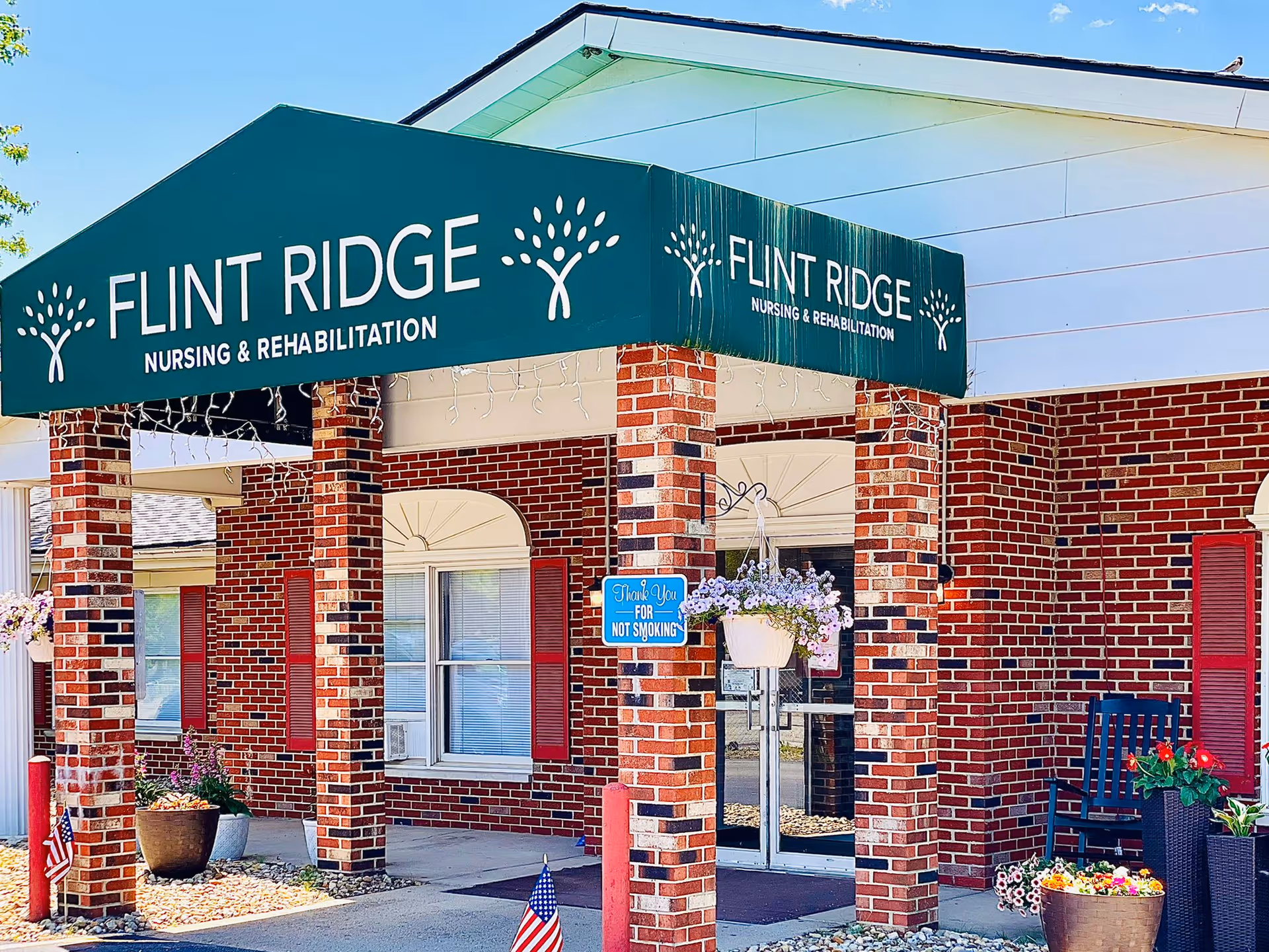 Entrance of Flint Ridge Nursing and Rehabilitation facility with a green awning displaying the facility name. The building has red brick walls, white trim, and red shutters. There are potted plants and American flags near the entrance, along with a blue sign that says 'Thank You For Not Smoking'.