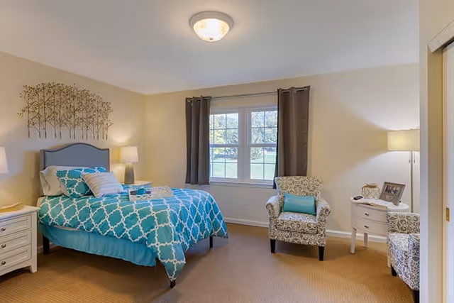 A cozy bedroom in Brookdale Northville featuring a bed with a blue and white patterned comforter, two matching pillows, and a folded towel on top. The room has beige walls, a window with dark curtains, a decorative metal wall art above the bed, two white nightstands with lamps, and two patterned armchairs with a small round white table between them holding a picture frame and a lamp.